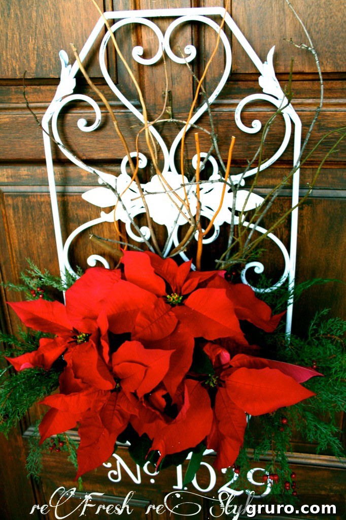 Elegant winter door decor with vibrant red poinsettias or similar winter flowers nestled in a decorative metal container, adorned with seasonal greenery.